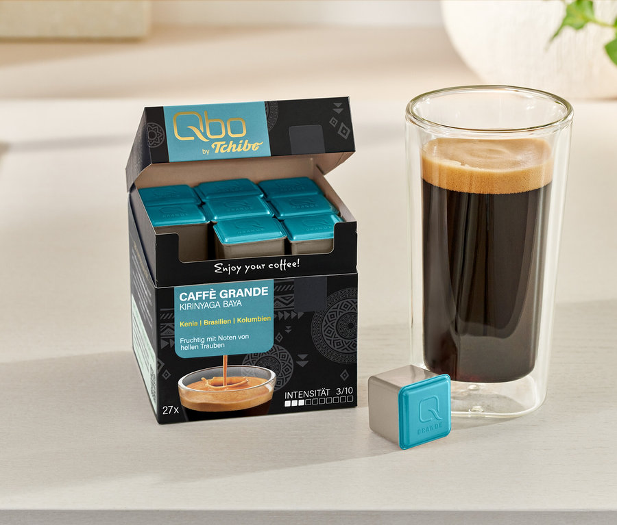 Une boîte ouverte de capsules Qbo Caffè Grande KIRINYAGA BAYA, un verre de café et une capsule individuelle sur une table.