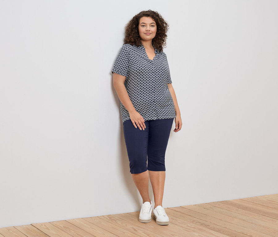 Une femme aux cheveux bouclés est appuyée contre un mur et porte un chemisier à motifs bleus et blancs, un pantacourt bleu et des baskets blanches.