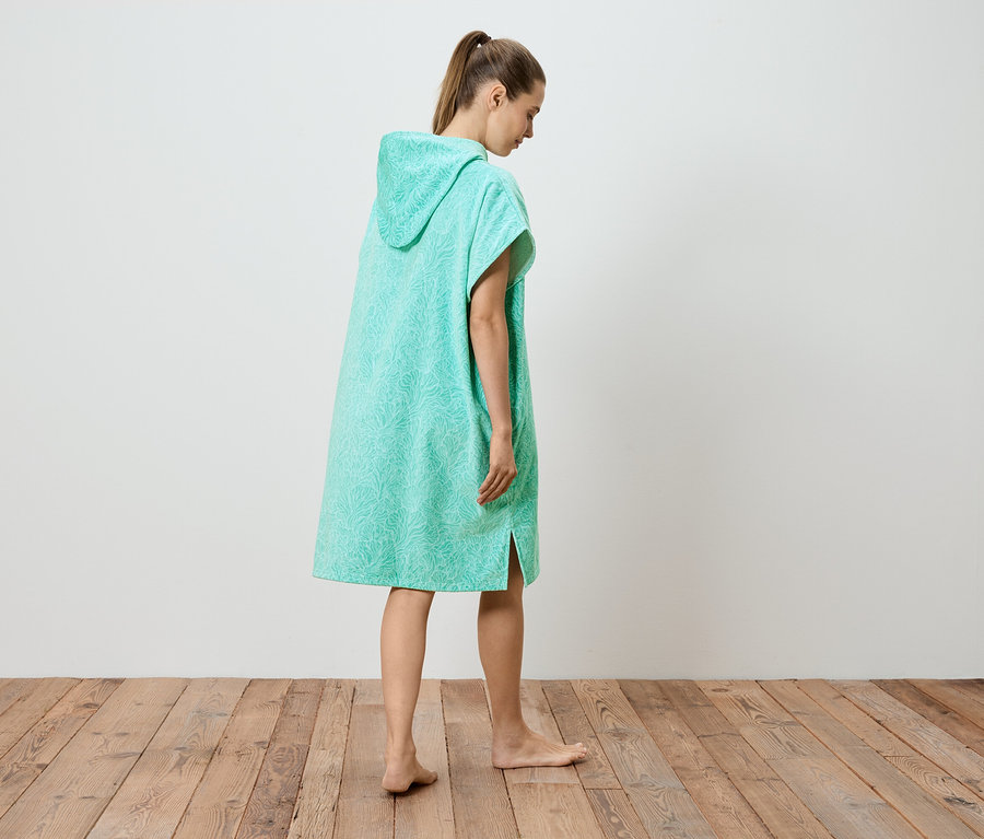 Une femme se tient pieds nus sur un plancher en bois, portant un poncho turquoise à capuche avec un motif.