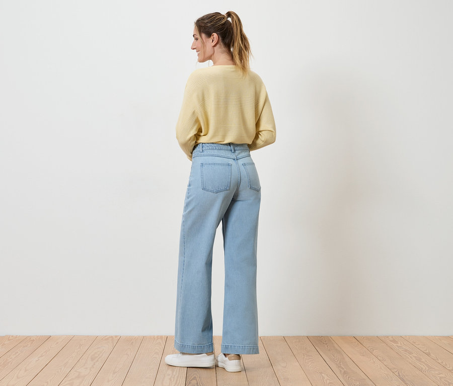 Une femme avec une queue de cheval porte un pull jaune clair, un jean large bleu clair et des baskets blanches, vue de dos.