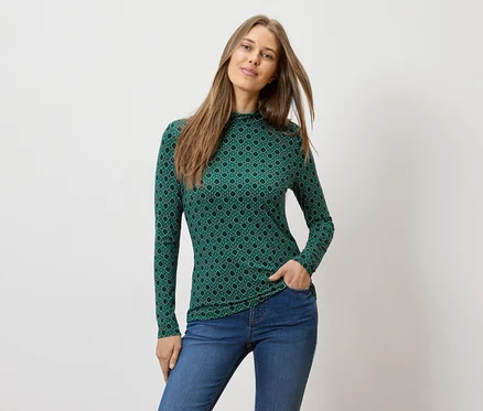 Une femme, une main dans la poche, porte un haut vert à manches longues avec un motif géométrique et un jean.