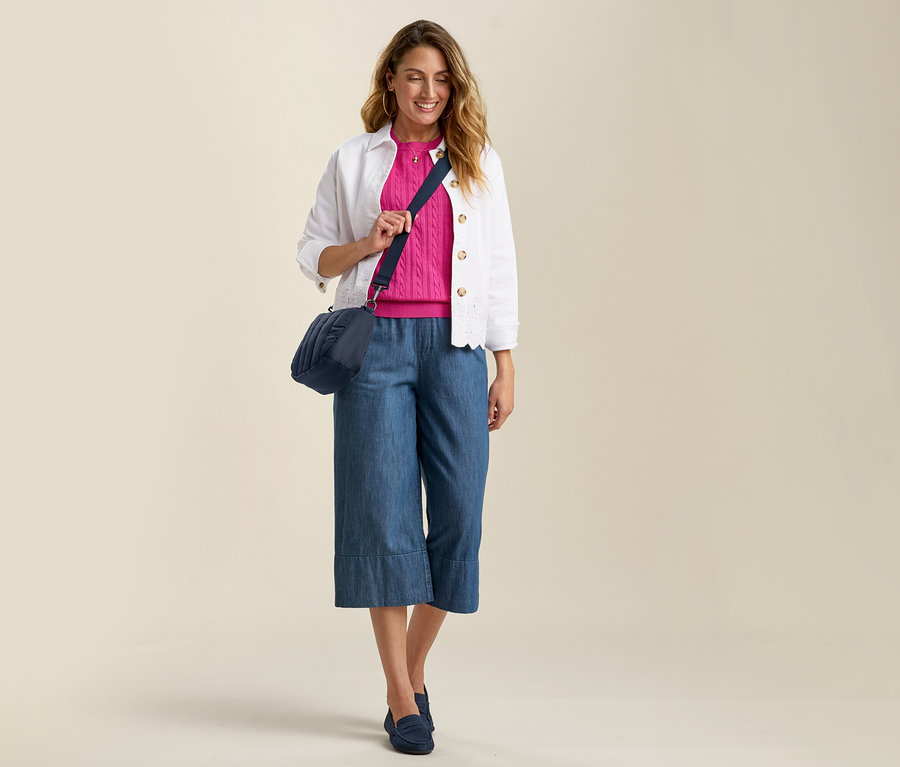 Femme portant une culotte de jean, un pull rose et une veste en jean avec broderie ajourée. Sac bandoulière bleu marine et mocassins assortis.