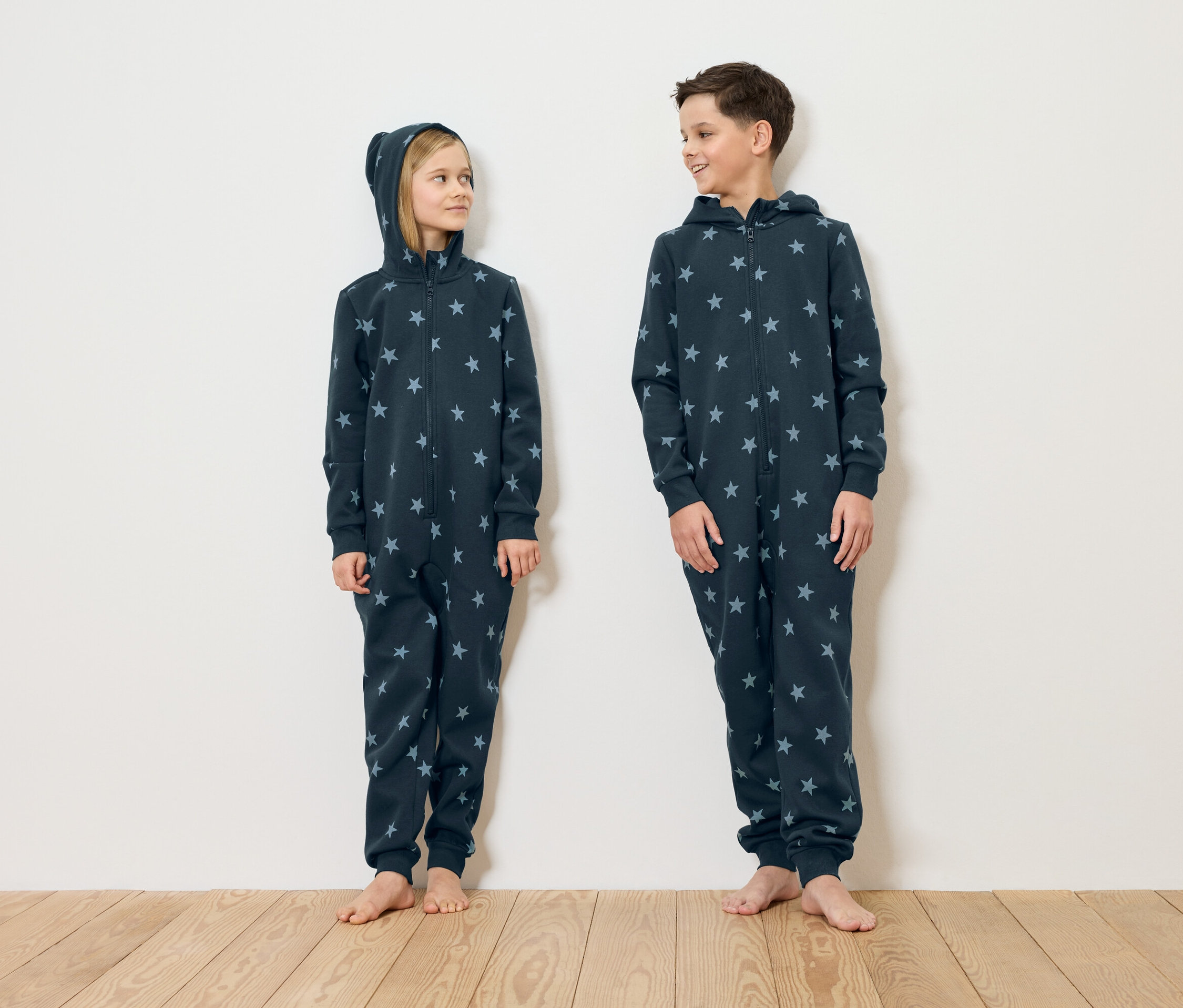 Une fille et un garçon debout pieds nus devant un mur blanc portant des pyjamas bleu marine à motifs étoilés.