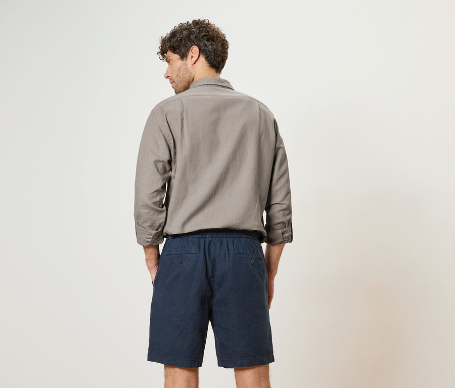 Un homme aux cheveux bouclés se tient dos à la caméra, portant une chemise grise et un short bleu.