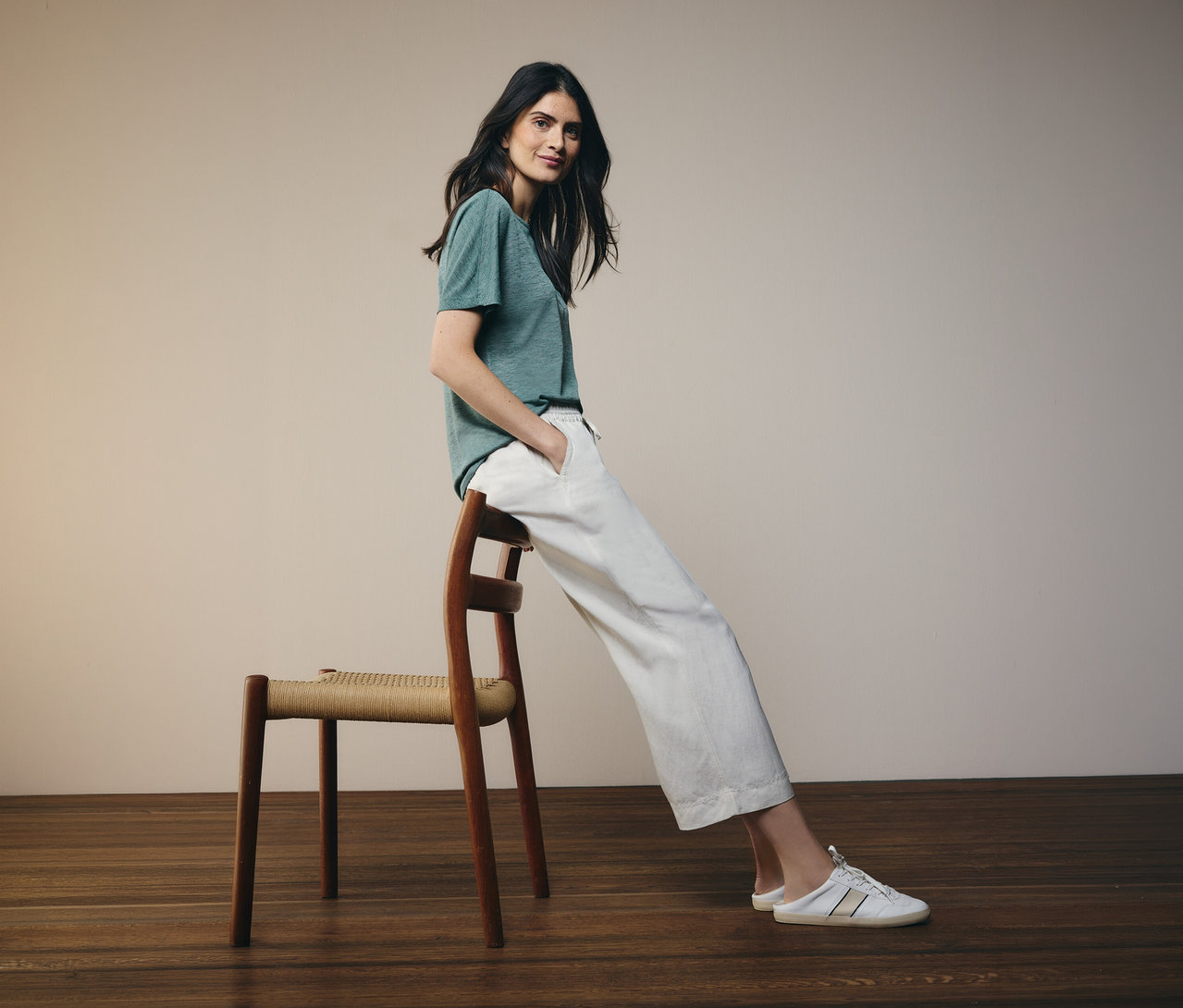 Une femme portant un t-shirt vert et un pantalon blanc est assise sur une chaise en bois avec une assise tressée sur un plancher en bois.