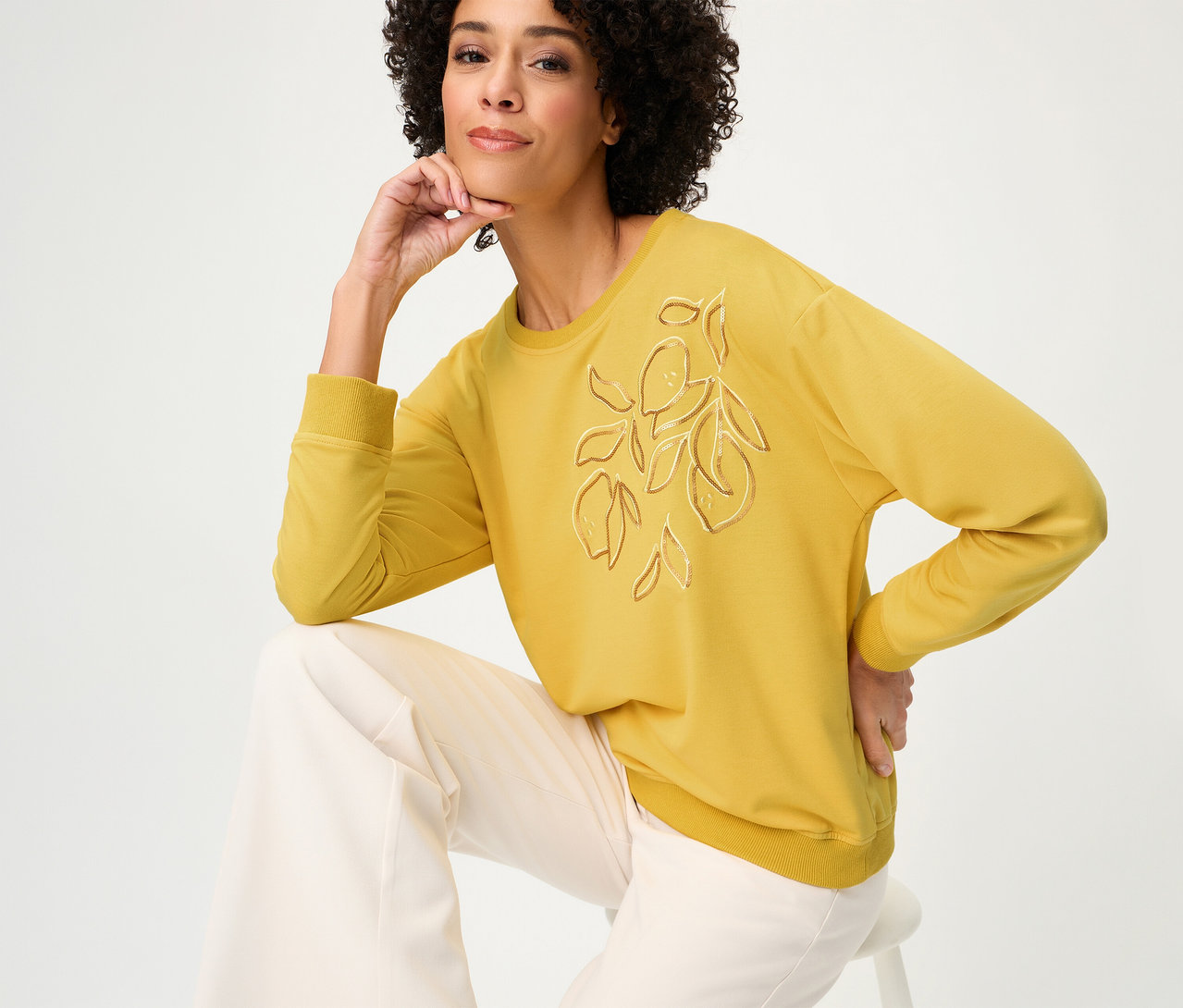 Femme assise sur un tabouret portant un sweat-shirt Olsen manches longues jaune avec motif citron et un pantalon ample de couleur claire.