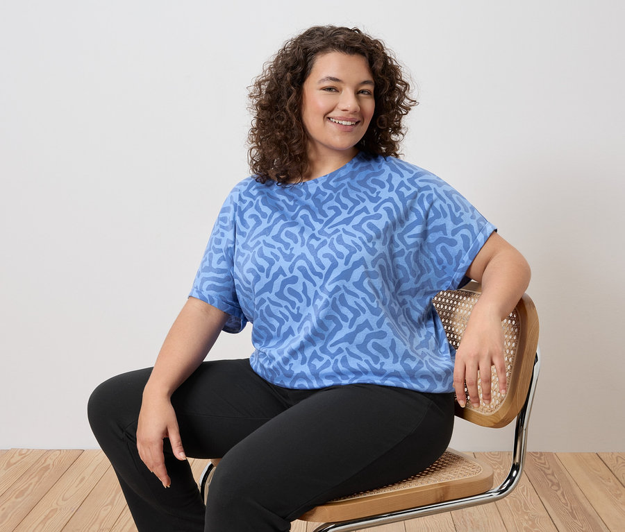 Une femme aux cheveux bouclés est assise sur une chaise, portant un t-shirt bleu à motifs et un pantalon noir.