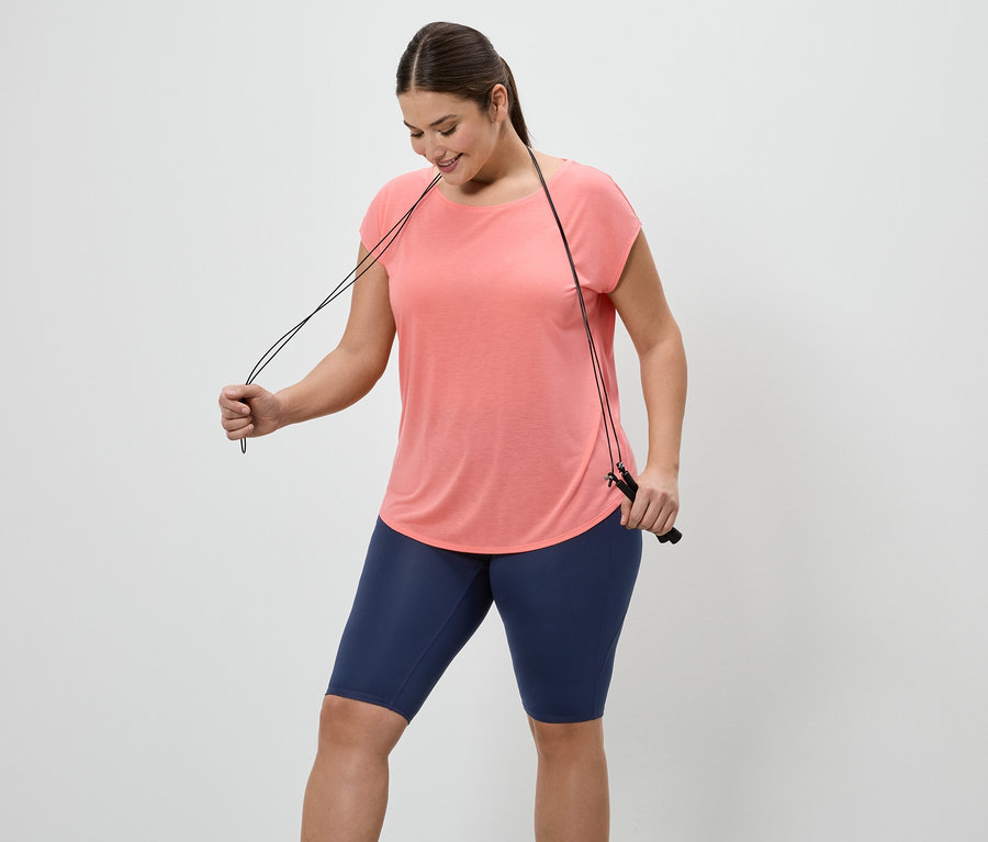 Une femme portant un t-shirt couleur saumon et un short de cycliste bleu tient une corde à sauter sur fond blanc.