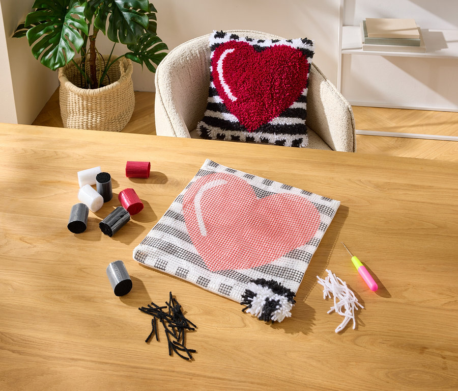 Un coussin en forme de cœur est posé sur une chaise. Sur une table, on trouve une housse de coussin inachevée, des rouleaux de fil, des morceaux de fil et un crochet.
