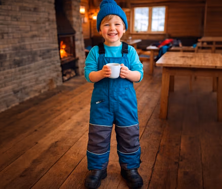 Un jeune garçon souriant portant un bonnet tricoté bleu, un t-shirt à manches longues bleu et une salopette de neige bleue et grise se tient sur un plancher en bois, tenant une tasse blanche. Une cheminée avec un feu est visible à sa gauche, et des tables et bancs en bois sont en arrière-plan.