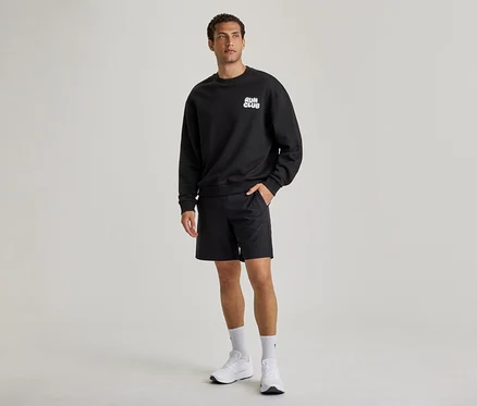 Un homme porte un sweatshirt noir avec l'inscription « RUN CLUB », un short noir et des baskets blanches. Sa main gauche est dans la poche de son short.
