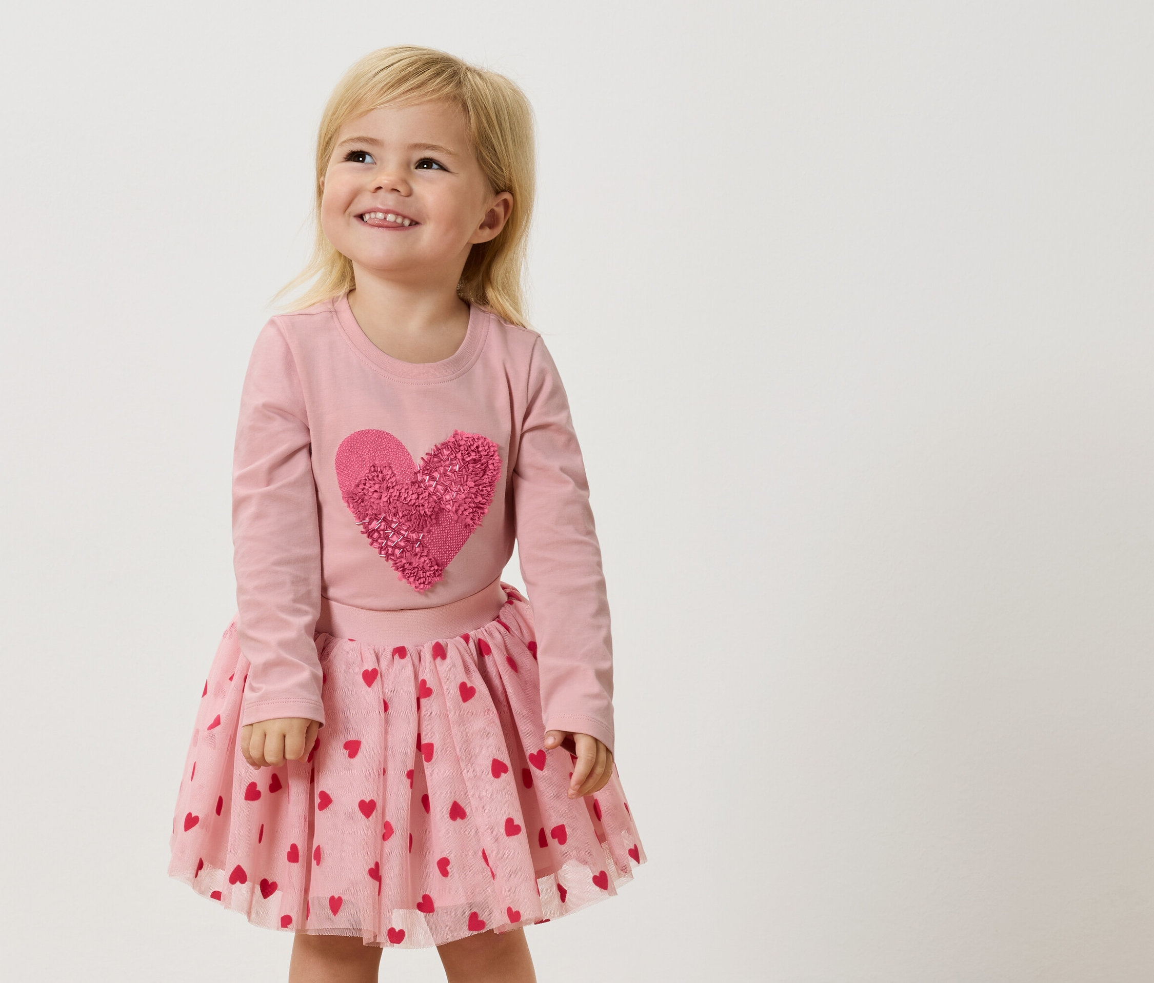 Une petite fille blonde porte un t-shirt rose à manches longues avec un grand cœur rose et une jupe en tulle rose avec des cœurs rouges.