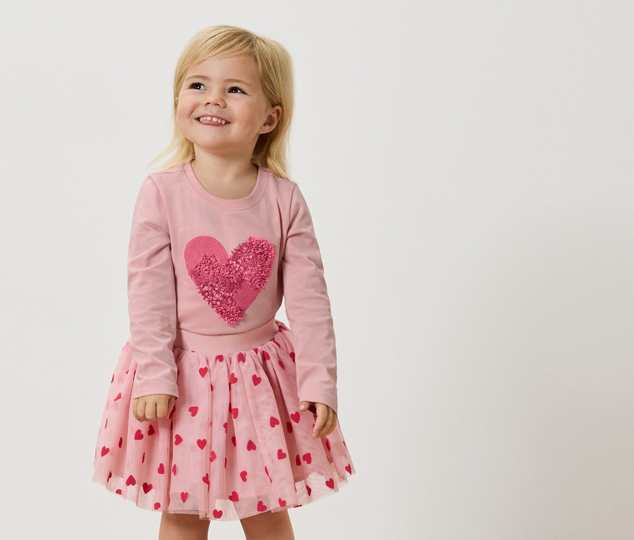 Une petite fille blonde porte un t-shirt rose à manches longues avec un grand cœur rose et une jupe en tulle rose avec des cœurs rouges.