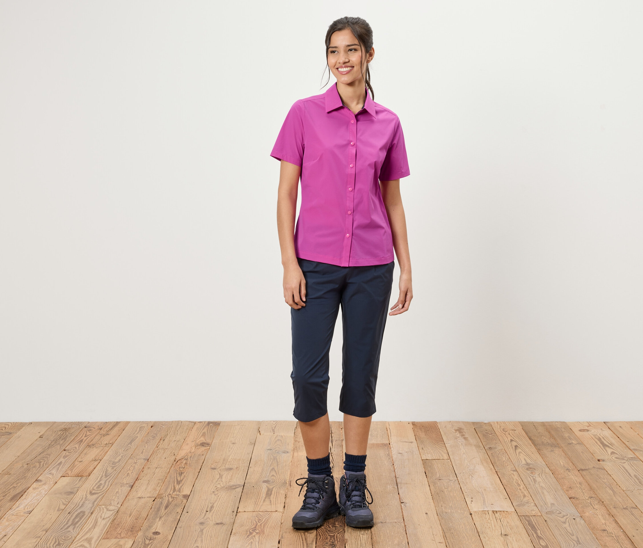 Une femme se tient devant un mur blanc. Elle porte une chemise rose, un pantacourt bleu et des chaussures de randonnée.