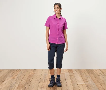 Une femme se tient devant un mur blanc. Elle porte une chemise rose, un pantacourt bleu et des chaussures de randonnée.