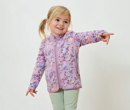 Une fillette souriante aux tresses blondes, vêtue d'une veste en polaire violette à motifs floraux et d'un pantalon vert clair, pointe du doigt.