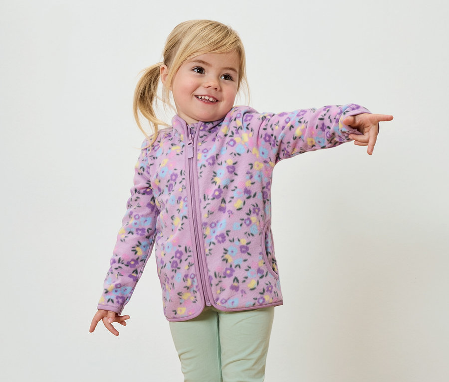 Une fillette souriante aux tresses blondes, vêtue d'une veste en polaire violette à motifs floraux et d'un pantalon vert clair, pointe du doigt.