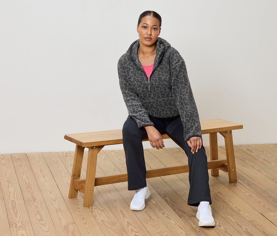 Une femme est assise sur un banc en bois et porte un pull en polaire gris à motif léopard, un haut rose, un pantalon foncé et des baskets blanches.