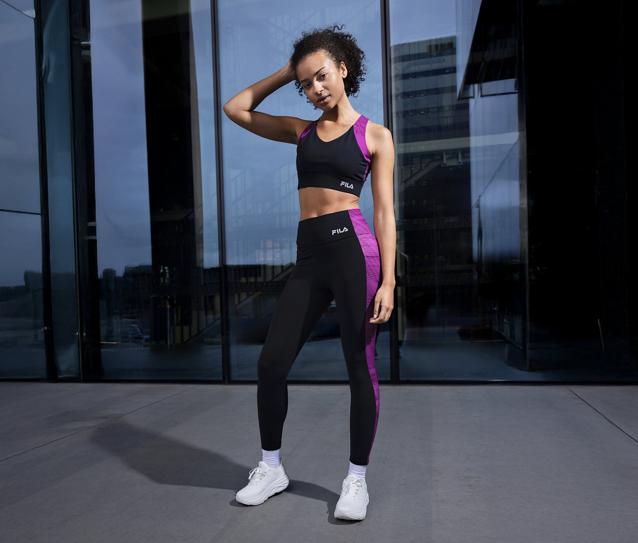 Femme en soutien-gorge de sport noir et leggings avec des bandes violettes posant devant un immeuble.