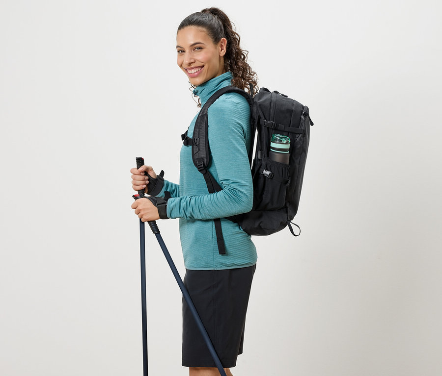 Femme portant un col roulé turquoise, un sac à dos noir et tenant des bâtons de randonnée.