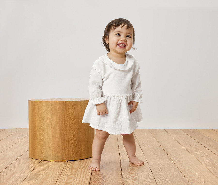 Un jeune enfant souriant et pieds nus se tient sur un plancher en bois à côté d'un tabouret cylindrique, portant une robe blanche.