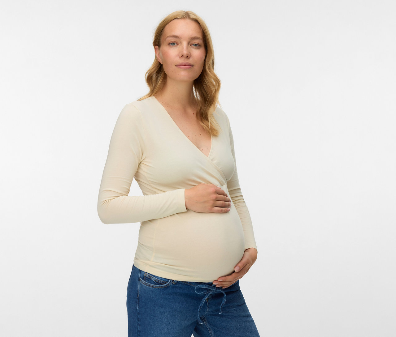 Femme enceinte portant un haut de grossesse beige MAMALICIOUS «
MLKERRIE » en lot de 2 et un jean, tenant son ventre.