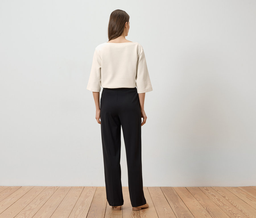 Femme vue de dos portant un haut crème et un pantalon noir sur un plancher en bois devant un mur blanc.