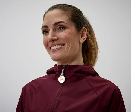 Une femme souriante avec une queue de cheval porte une veste à capuche bordeaux avec un pendentif rond et lumineux sur la fermeture éclair.