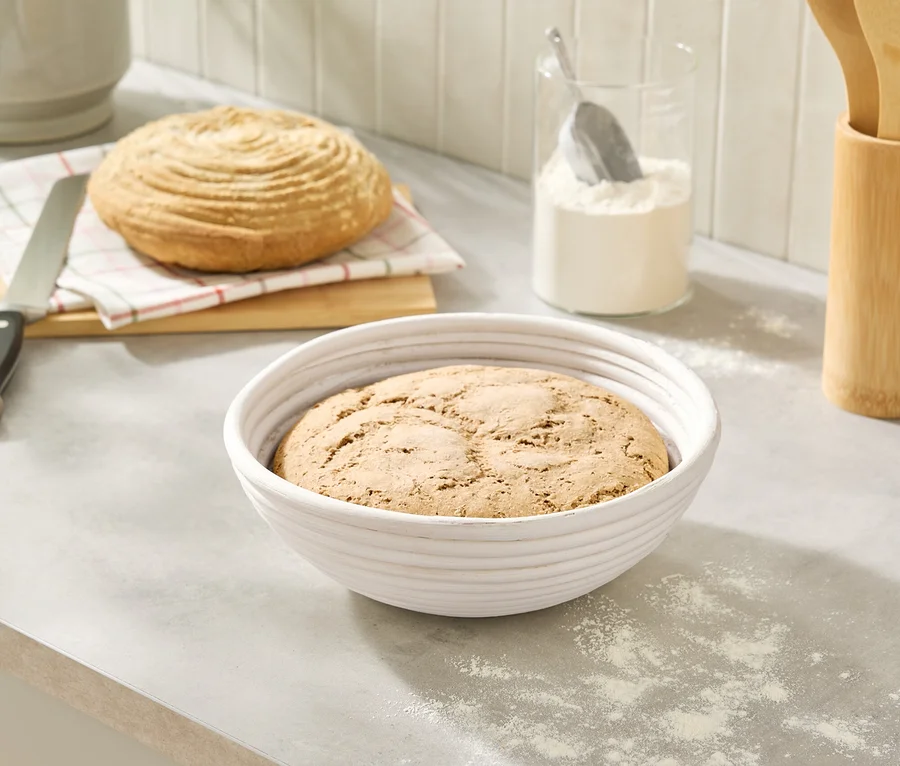 Sur un comptoir de cuisine, un pain dans un banneton blanc et un pain sur une planche à côté d'un couteau et d'un pot de farine.