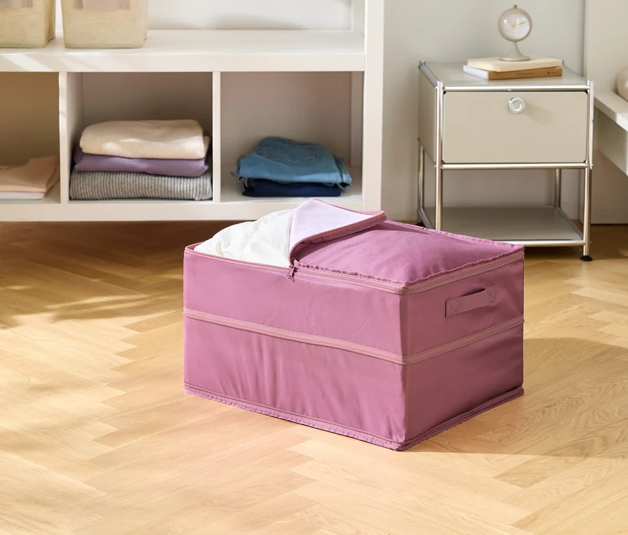 Un conteneur en tissu rose est posé sur un plancher en bois, partiellement rempli de linge blanc. On aperçoit une étagère et une table de chevet en arrière-plan.
