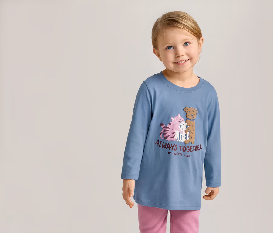 Fillette souriante portant un T-shirt bleu à manches longues avec un motif de chats et de chiens et l'inscription « Always Together » et un pantalon rose.
