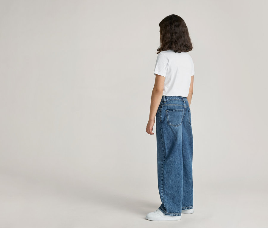 Vue arrière d'une personne portant un t-shirt blanc, un jean bleu et des chaussures blanches sur un fond clair.