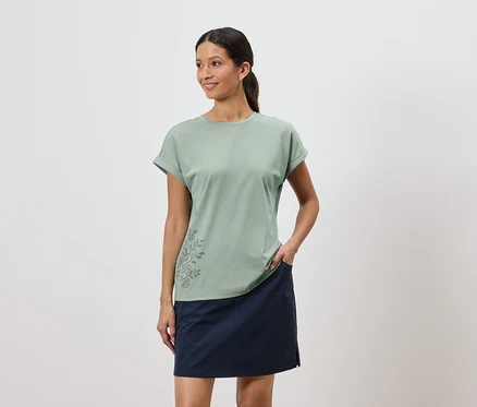 Femme portant un t-shirt vert clair avec un motif floral et une jupe bleue, une main dans la poche de la jupe.