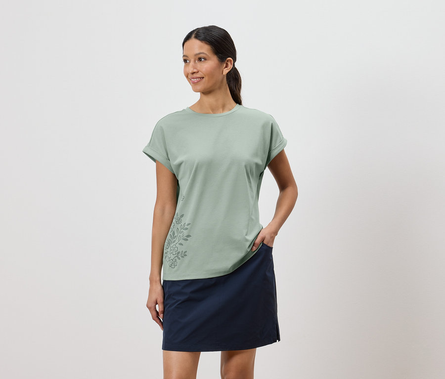 Femme portant un t-shirt vert clair avec un motif floral et une jupe bleue, une main dans la poche de la jupe.