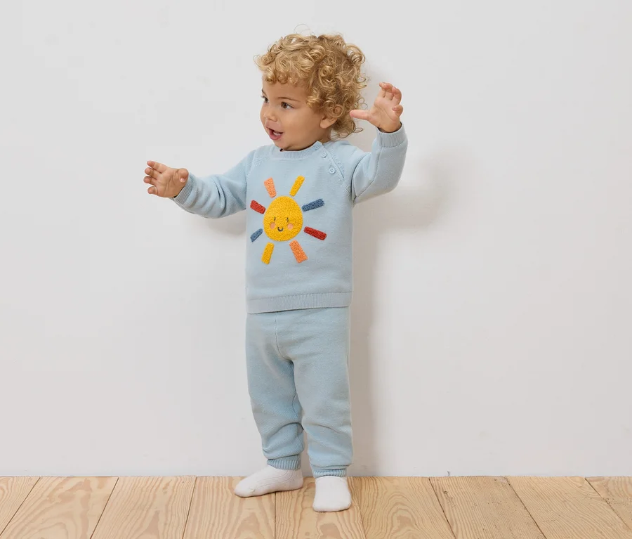 Un jeune enfant aux cheveux bouclés se tient devant un mur blanc, vêtu d'un pull et d'un pantalon bleu clair avec un motif de soleil.