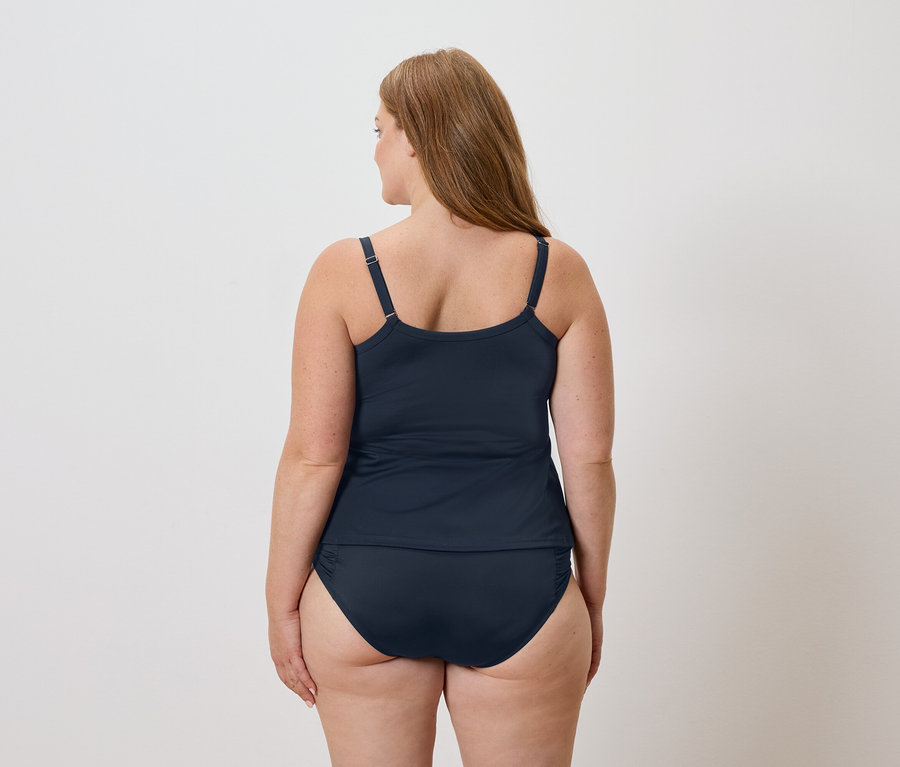 Vue arrière d'une femme aux longs cheveux bruns portant un haut de tankini bleu marine et un bas de maillot de bain sur fond blanc.