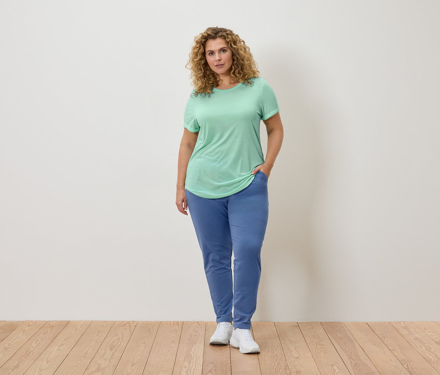 Femme aux cheveux bouclés portant un t-shirt vert clair et un pantalon de jogging bleu, debout sur un plancher en bois.