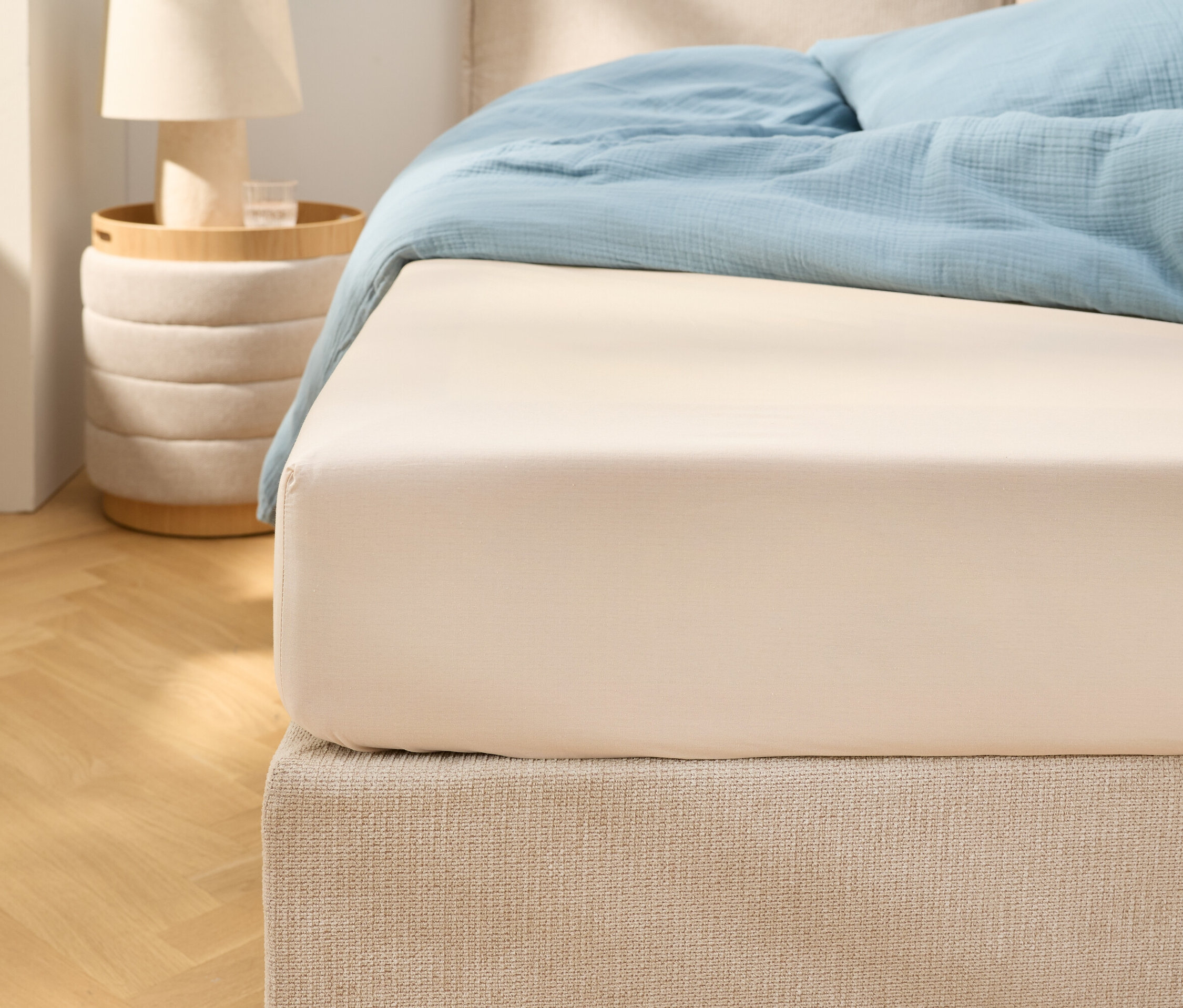 Gros plan d'un drap-housse beige sur un lit avec une literie bleu clair. Une table de chevet avec une lampe se trouve à côté.