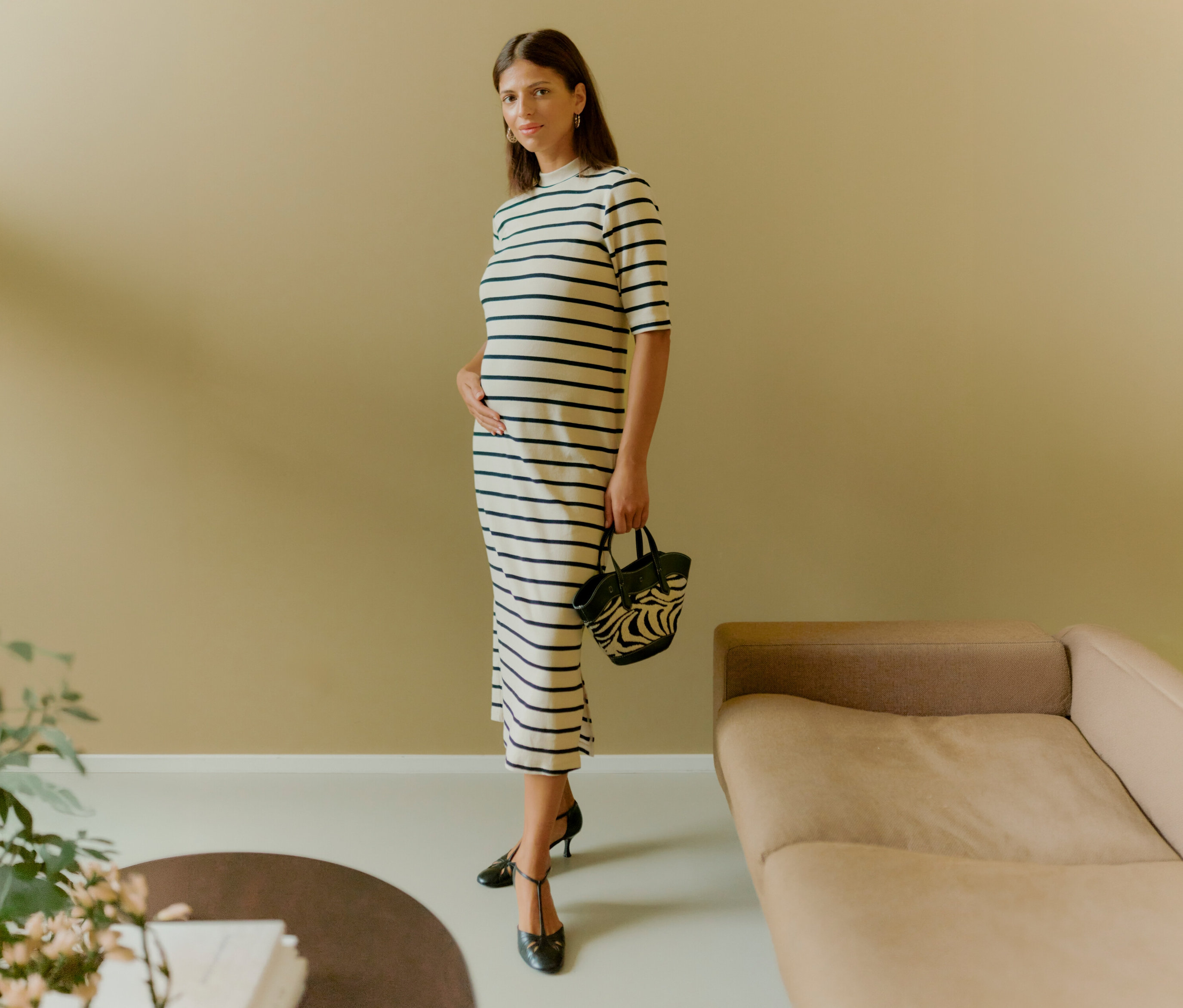 Une femme porte une robe MAMALICIOUS « MLLANE » rayée, une main sur le ventre, et tient un sac à main. Elle porte des chaussures noires.