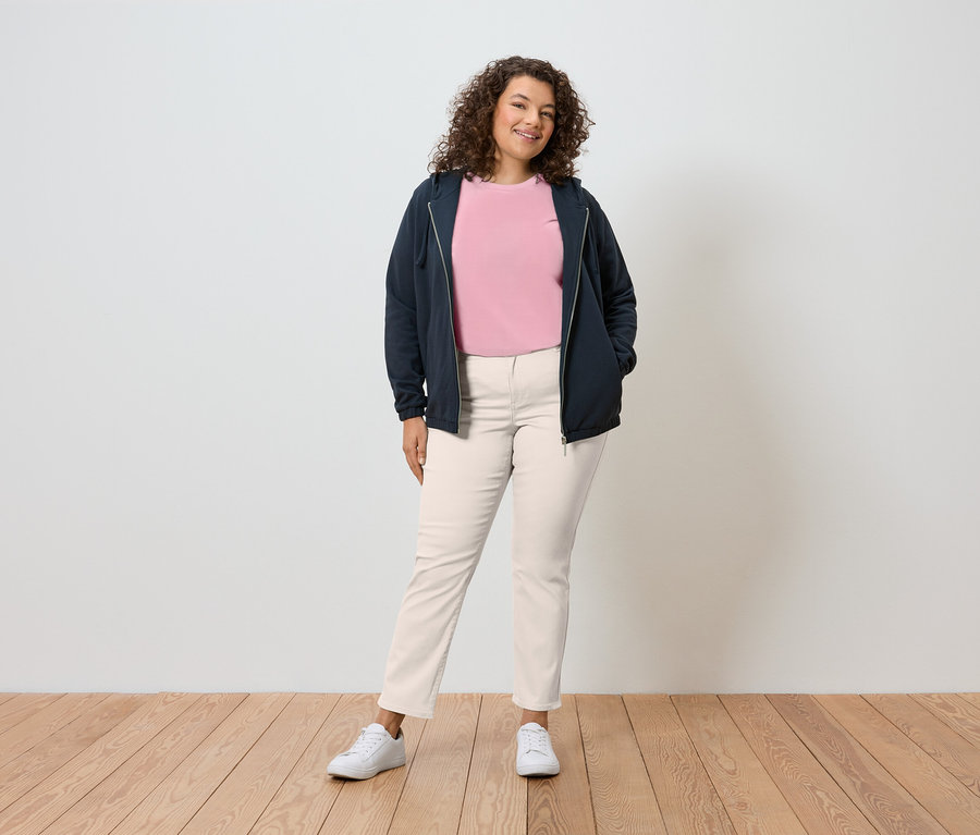 Une femme aux cheveux bouclés se tient devant un mur, portant un sweat à capuche bleu marine, un t-shirt rose, un pantalon crème et des baskets blanches.