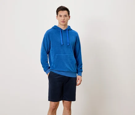 Un homme se tient debout, une main dans la poche de son pantalon, devant un fond neutre. Il porte un sweat à capuche bleu et un short court bleu marine.