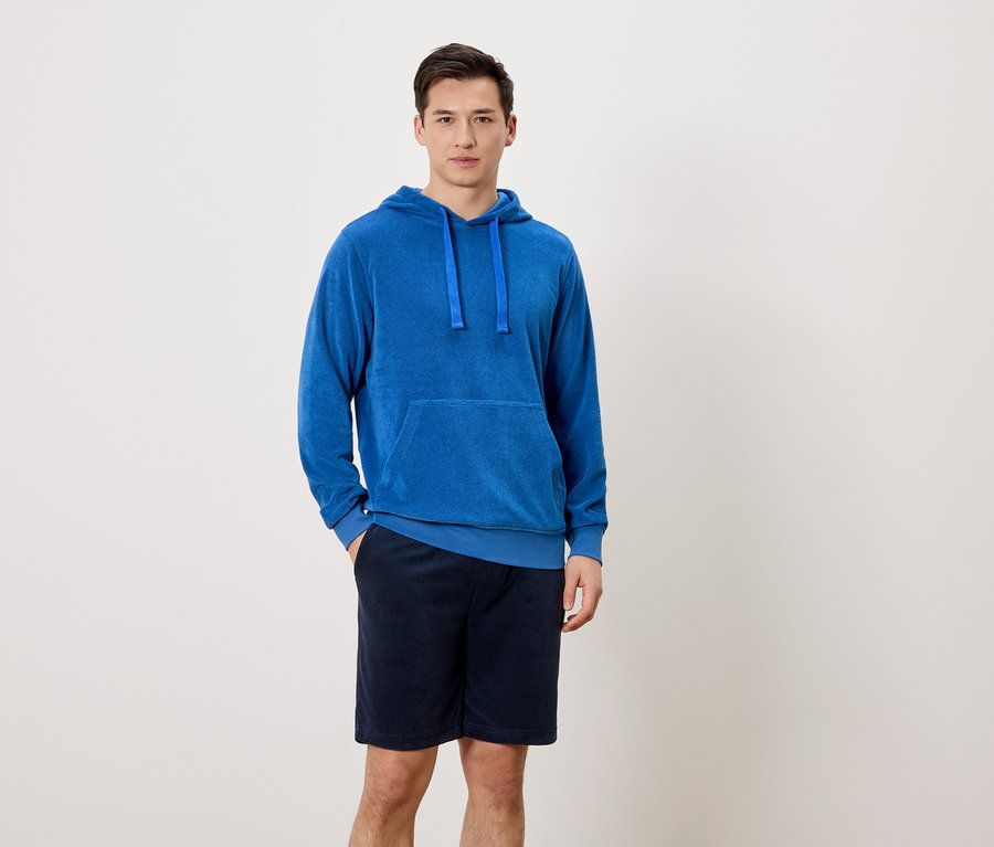 Un homme se tient debout, une main dans la poche de son pantalon, devant un fond neutre. Il porte un sweat à capuche bleu et un short court bleu marine.