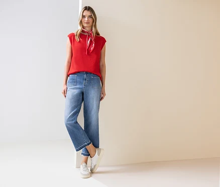 Une femme se tient debout et porte un haut rouge, un foulard, un jean et des baskets.