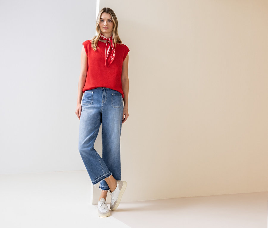 Une femme se tient debout et porte un haut rouge, un foulard, un jean et des baskets.