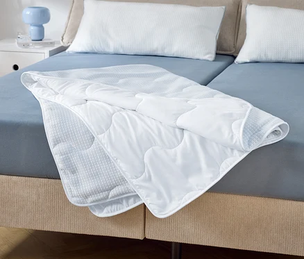 Sur un lit double avec des draps bleus se trouve une couette blanche matelassée partiellement pliée. Deux oreillers blancs sont posés à la tête de lit.