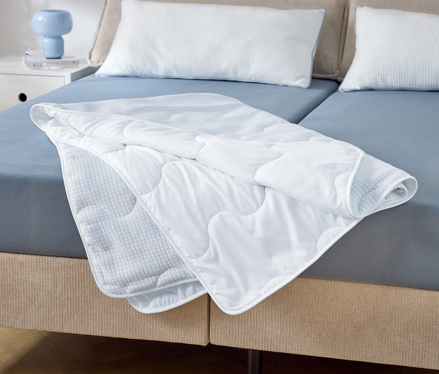 Sur un lit double avec des draps bleus se trouve une couette blanche matelassée partiellement pliée. Deux oreillers blancs sont posés à la tête de lit.