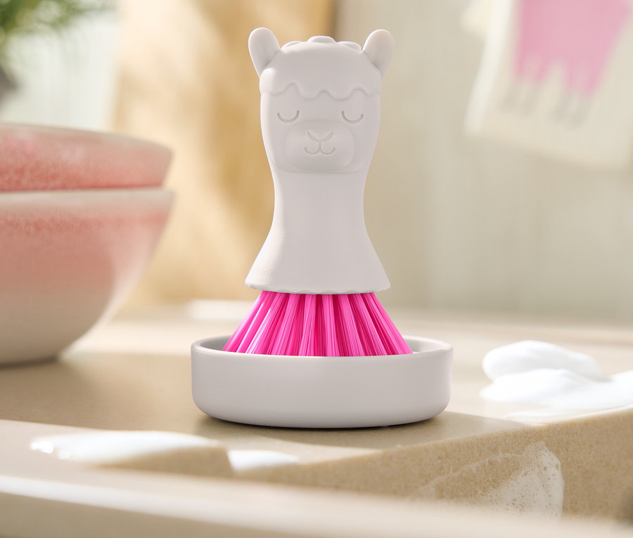 Une brosse blanche à poils roses en forme de lama est posée dans un récipient blanc sur un évier avec des résidus de savon.