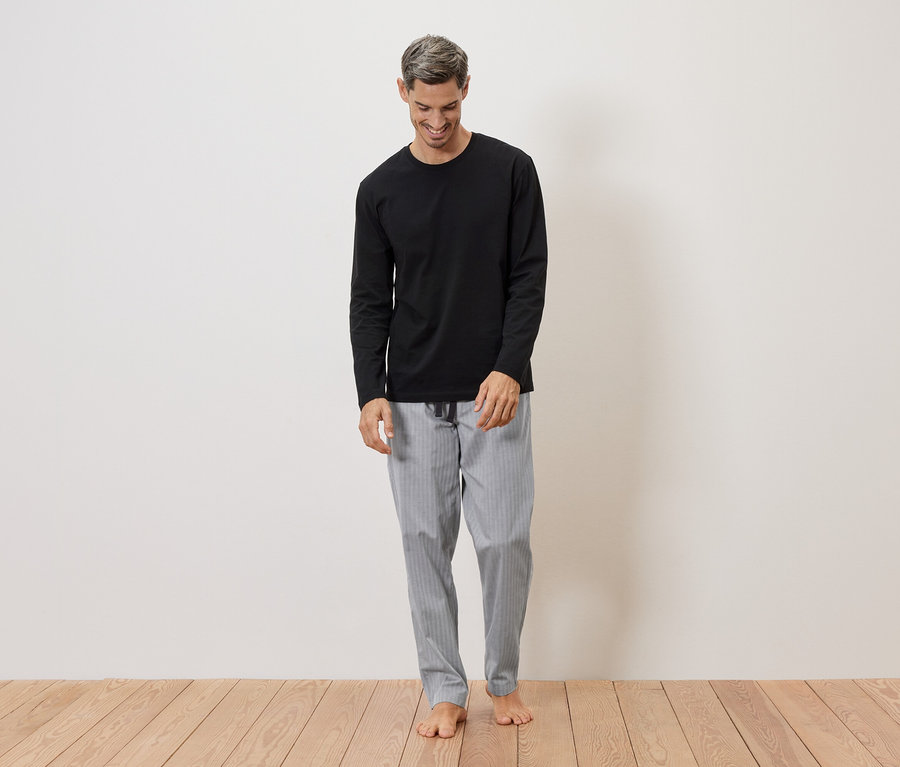 Un homme souriant aux cheveux gris porte un t-shirt noir à manches longues et un pantalon de pyjama gris rayé, pieds nus sur un plancher en bois.