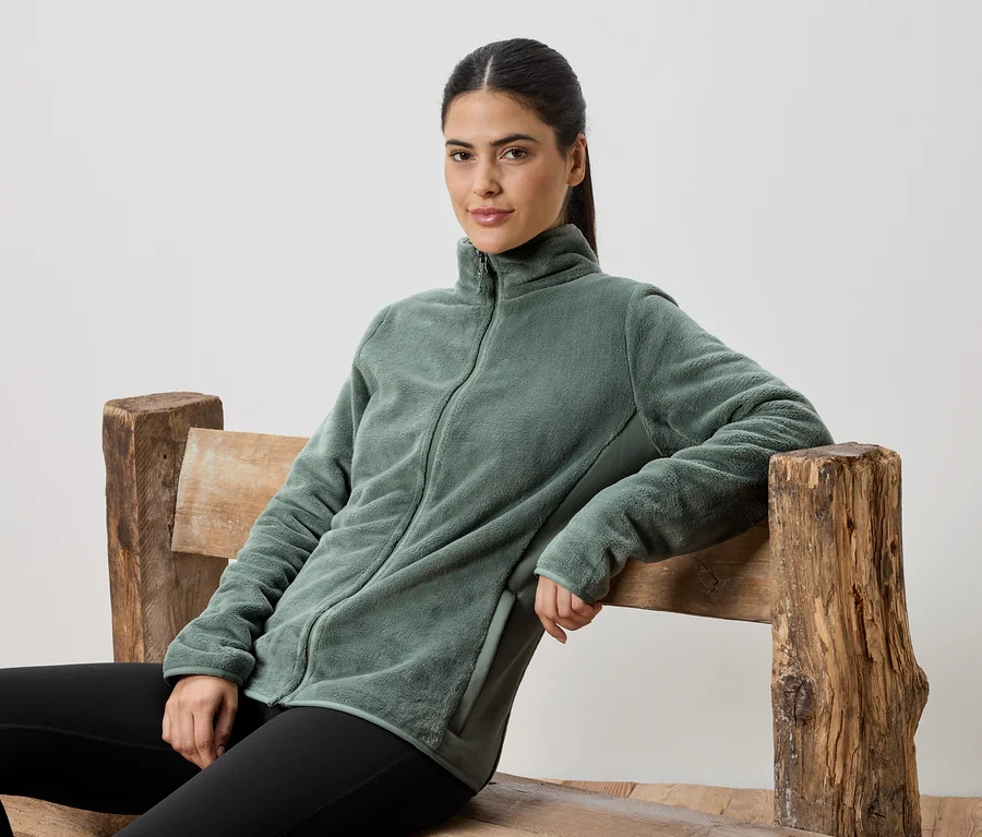 Une femme assise sur un banc en bois porte une veste en polaire verte et un legging noir.