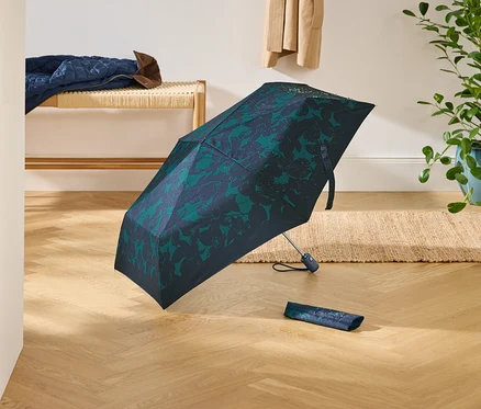 Parapluie bleu marine ouvert avec motif floral vert et son étui sur un plancher en bois. À l'arrière-plan, banc, plante et vêtements.